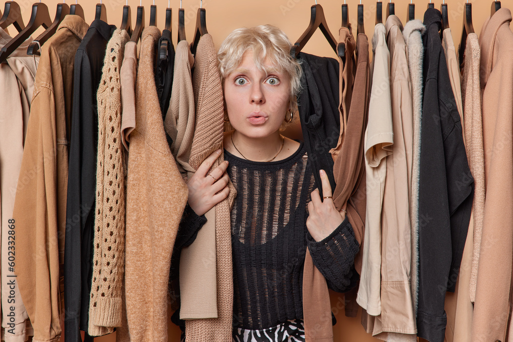 Shocked blonde young woman stands near clothes hang on rail feels excited hears something unbelevable chooses outfit to wear from her wardrobe buys new spring trends. Women and fashion concept
