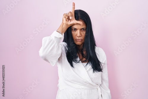 Mature hispanic woman standing over pink background making fun of people with fingers on forehead doing loser gesture mocking and insulting.