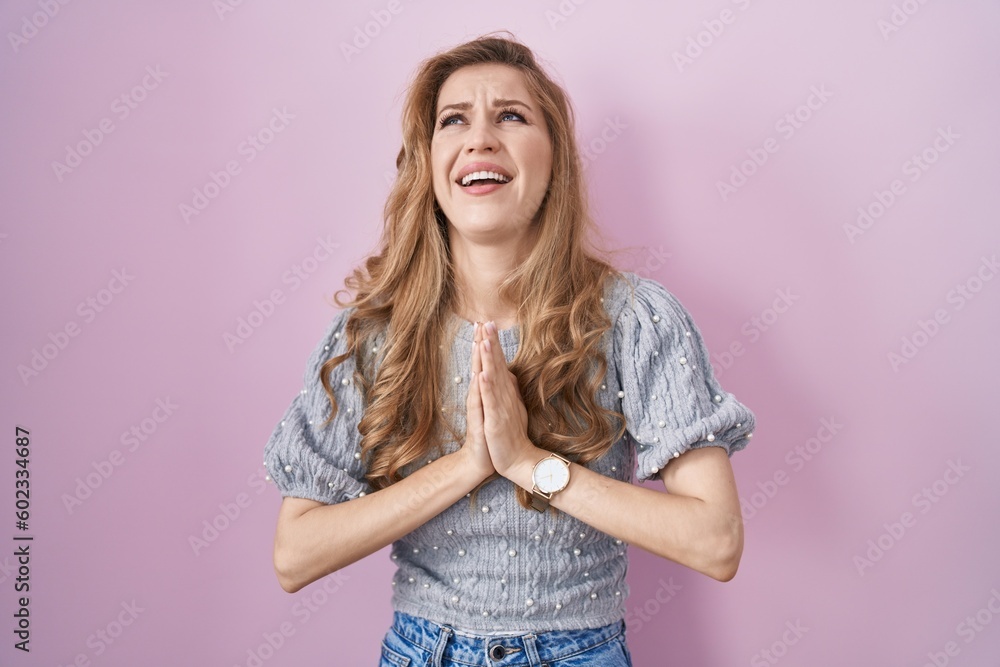 Beautiful blonde woman standing over pink background begging and praying with hands together with hope expression on face very emotional and worried. begging. Beautiful blonde woman standing over pink background begging and praying with hands together with hope expression on face very emotional and worried. begging.