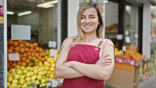 Young blonde woman shop assistant standing with arms crossed gesture at fruit store
