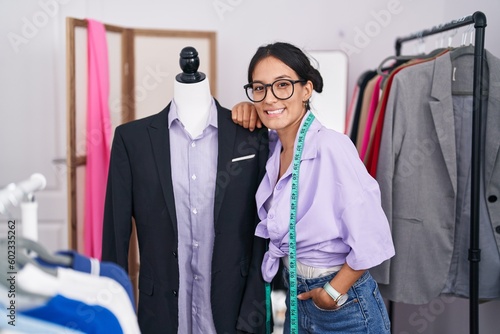 Young beautiful hispanic woman tailor smiling confident leaning on manikin at tailor shop