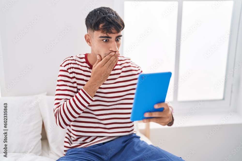 Young hispanic man using touchpad sitting on the bed covering mouth with hand, shocked and afraid for mistake. surprised expression Young hispanic man using touchpad sitting on the bed covering mouth with hand, shocked and afraid for mistake. surprised expression