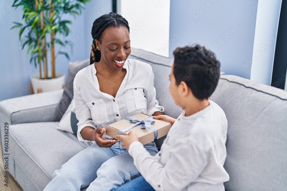 Fototapeta premium African american mother and son surprise with birthday gift sitting on sofa at home