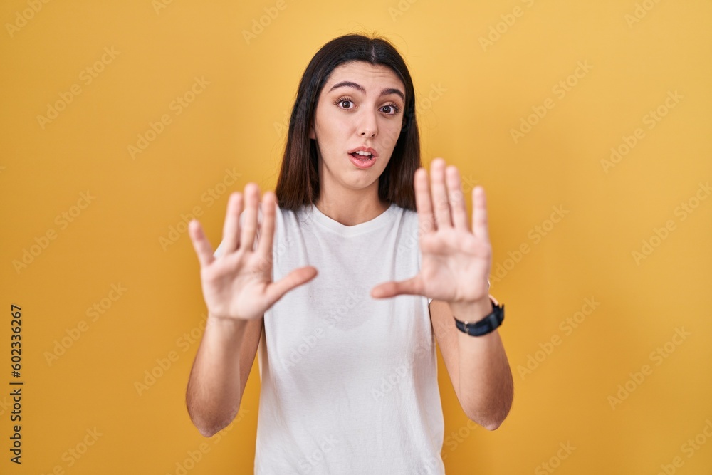Young beautiful woman standing over yellow background afraid and terrified with fear expression stop gesture with hands, shouting in shock. panic concept. Young beautiful woman standing over yellow background afraid and terrified with fear expression stop gesture with hands, shouting in shock. panic concept.