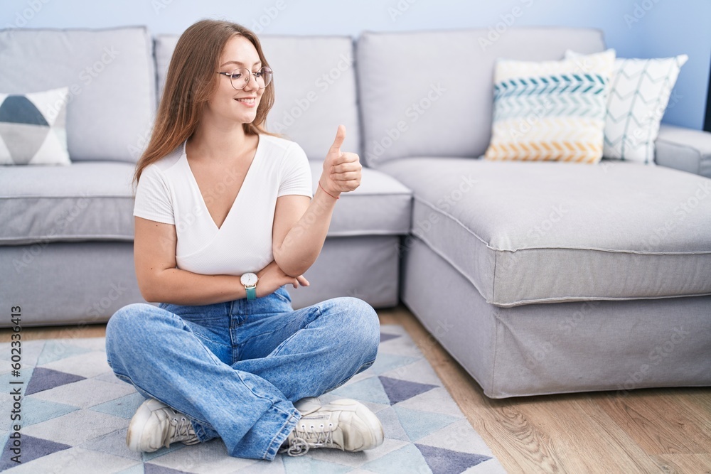 Young caucasian woman sitting on the floor at the living room looking proud, smiling doing thumbs up gesture to the side Young caucasian woman sitting on the floor at the living room looking proud, smiling doing thumbs up gesture to the side