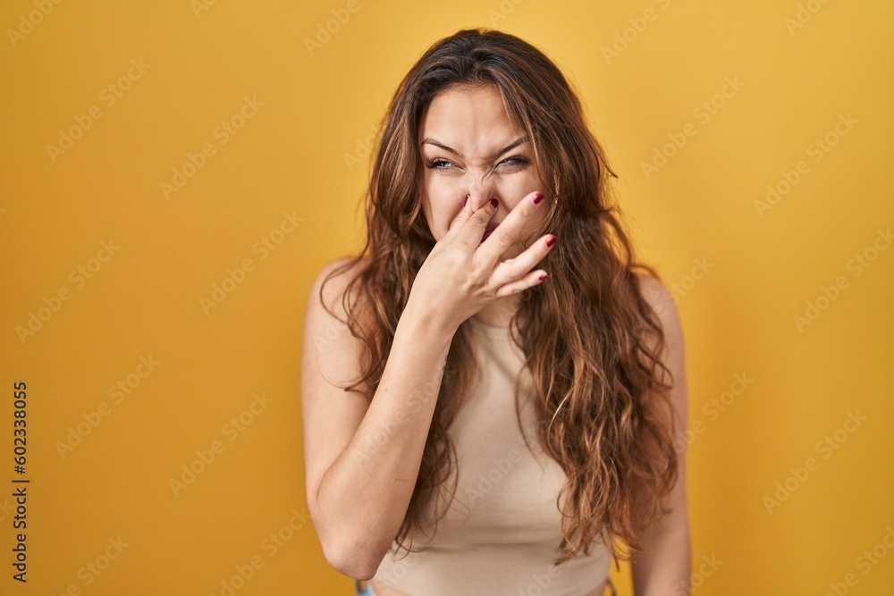 Young hispanic woman standing over yellow background smelling something stinky and disgusting, intolerable smell, holding breath with fingers on nose. bad smell Young hispanic woman standing over yellow background smelling something stinky and disgusting, intolerable smell, holding breath with fingers on nose. bad smell