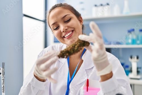 Young beautiful hispanic woman scientist smiling confident holding marijuana at laboratory