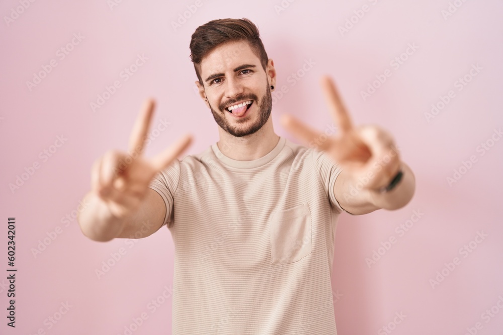 Hispanic man with beard standing over pink background smiling with tongue out showing fingers of both hands doing victory sign. number two. Hispanic man with beard standing over pink background smiling with tongue out showing fingers of both hands doing victory sign. number two.