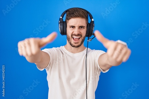 Hispanic man with beard listening to music wearing headphones approving doing positive gesture with hand, thumbs up smiling and happy for success. winner gesture.