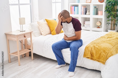 Young latin man suffering for stomach and head ache sitting on bed at bedroom