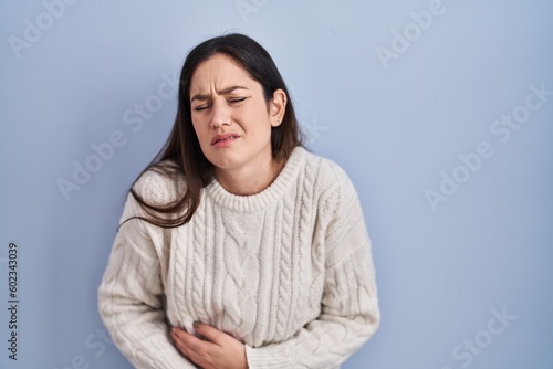 Young brunette woman standing over blue background with hand on stomach because indigestion, painful illness feeling unwell. ache concept.