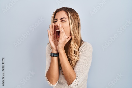 Young blonde woman standing over isolated background shouting angry out loud with hands over mouth