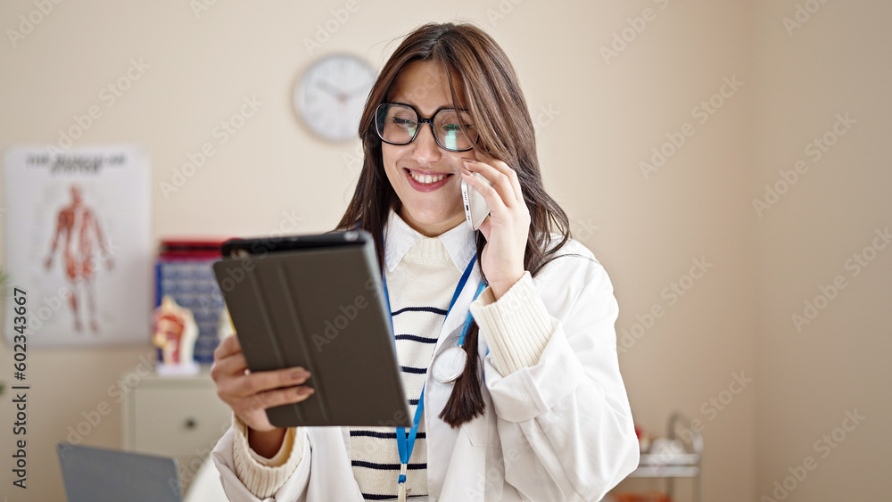 Young beautiful hispanic woman doctor using touchpad talking on smartphone at clinic Young beautiful hispanic woman doctor using touchpad talking on smartphone at clinic