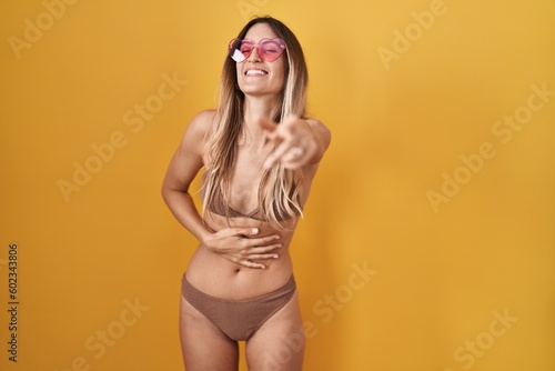 Young hispanic woman wearing bikini over yellow background laughing at you, pointing finger to the camera with hand over body, shame expression