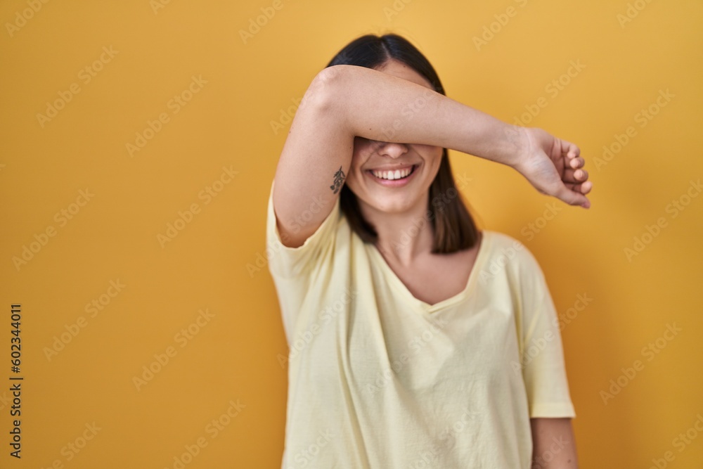 Hispanic girl wearing casual t shirt over yellow background covering eyes with arm smiling cheerful and funny. blind concept. Hispanic girl wearing casual t shirt over yellow background covering eyes with arm smiling cheerful and funny. blind concept.
