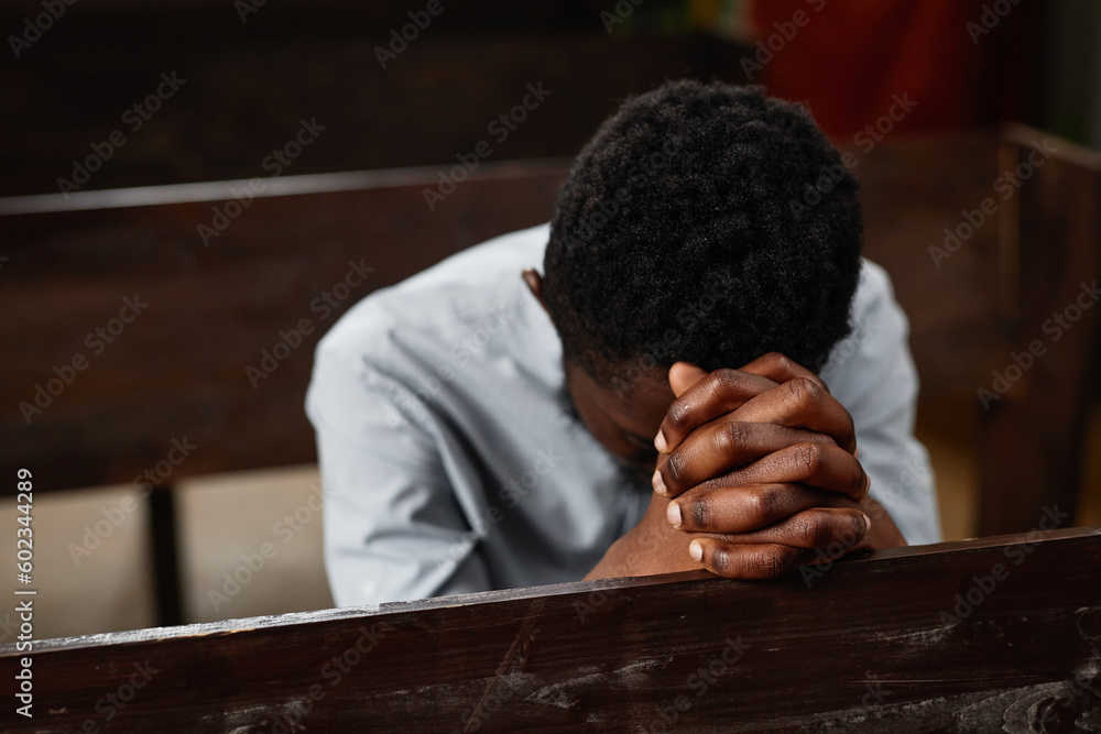Fototapeta premium Young African American man sitting on bench and praying in baptist church
