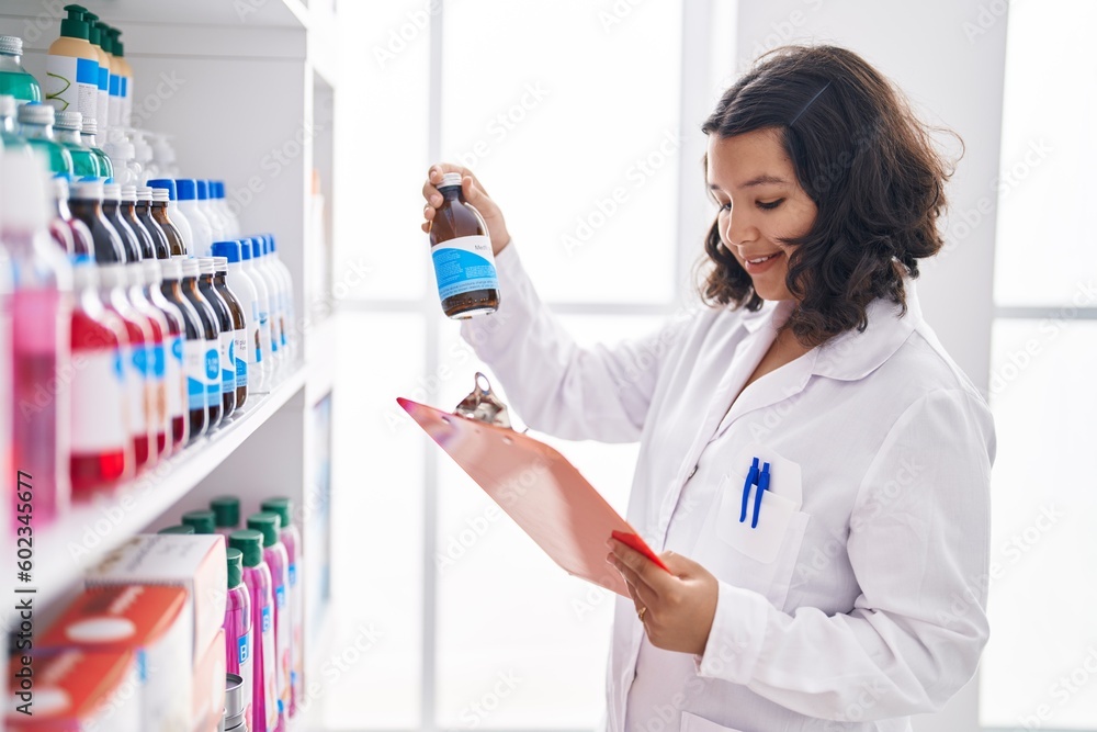 Young woman pharmacist holding medication bottle reading document at pharmacy