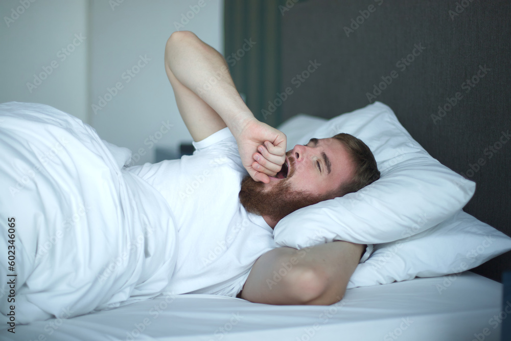 Young happy handsome sleepy man in the bed in bedroom at home in the morning lying under white blanket, enjoy resting, sleep well in night mask on face, eyes Young happy handsome sleepy man in the bed in bedroom at home in the morning lying under white blanket, enjoy resting, sleep well in night mask on face, eyes