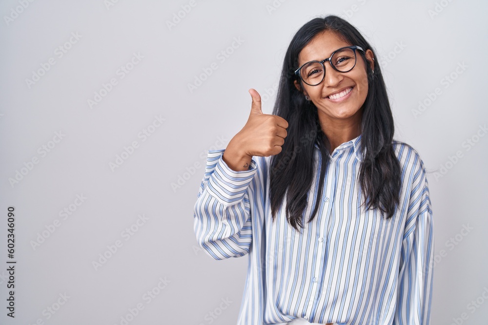 Young hispanic woman wearing glasses doing happy thumbs up gesture with hand. approving expression looking at the camera showing success. Young hispanic woman wearing glasses doing happy thumbs up gesture with hand. approving expression looking at the camera showing success.