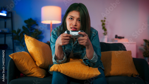 Excited beautiful Asian woman using joystick wireless controller enjoy playing fighting console video game to win highest victory. Girl Gamer sit on sofa in neon living room.