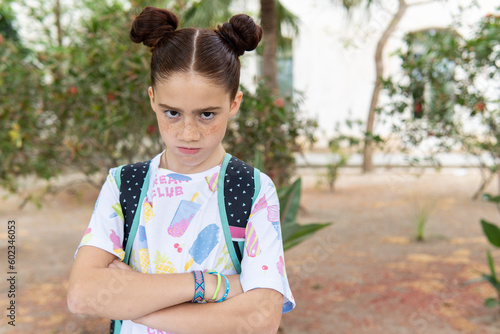 Portrait of a red haired girl with freckles looking at camera with her arms crossed and an angry face in a park, carrying a backpack.