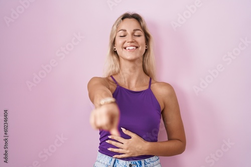 Young blonde woman standing over pink background laughing at you  pointing finger to the camera with hand over body  shame expression