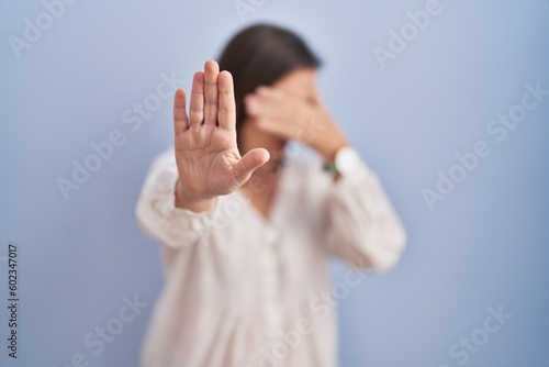 Middle age hispanic woman standing over blue background covering eyes with hands and doing stop gesture with sad and fear expression. embarrassed and negative concept.