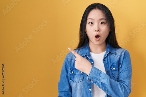 Young asian woman standing over yellow background surprised pointing with finger to the side, open mouth amazed expression.