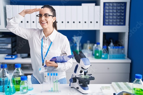 Young brunette woman working at scientist laboratory very happy and smiling looking far away with hand over head. searching concept.