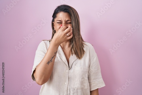 Blonde woman standing over pink background smelling something stinky and disgusting, intolerable smell, holding breath with fingers on nose. bad smell © Krakenimages.com