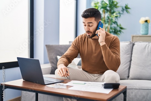 Young hispanic man accounting economy talking on the smartphone at home