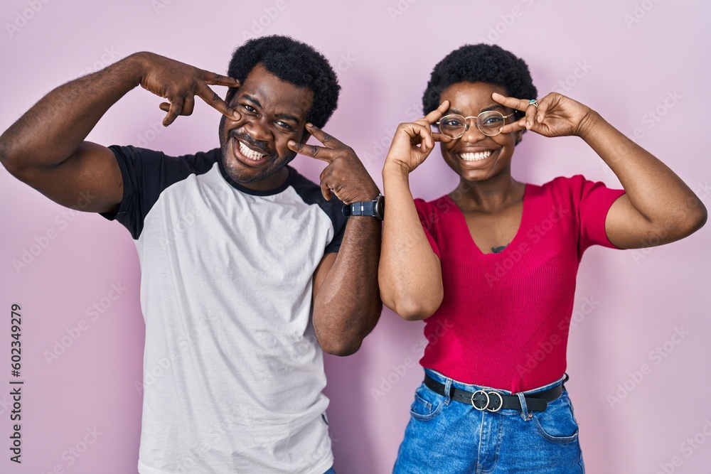 Young african american couple standing over pink background doing peace symbol with fingers over face, smiling cheerful showing victory Young african american couple standing over pink background doing peace symbol with fingers over face, smiling cheerful showing victory