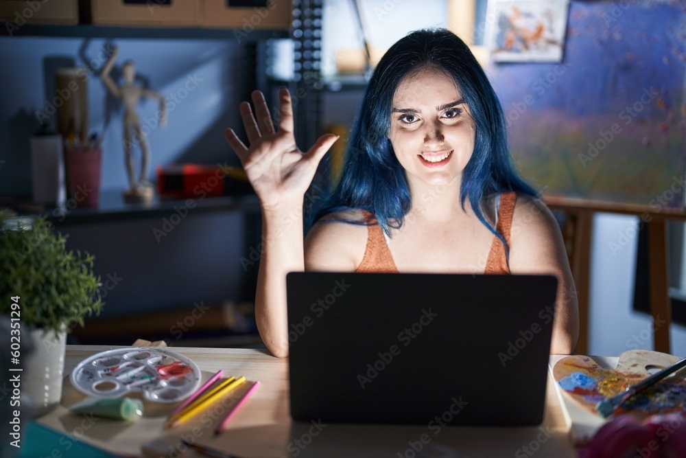 Young modern girl with blue hair sitting at art studio with laptop at night showing and pointing up with fingers number five while smiling confident and happy. Young modern girl with blue hair sitting at art studio with laptop at night showing and pointing up with fingers number five while smiling confident and happy.