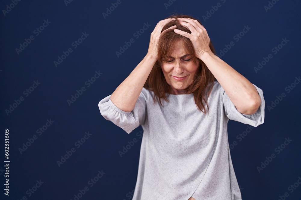Middle age woman standing over blue background suffering from headache desperate and stressed because pain and migraine. hands on head. Middle age woman standing over blue background suffering from headache desperate and stressed because pain and migraine. hands on head.