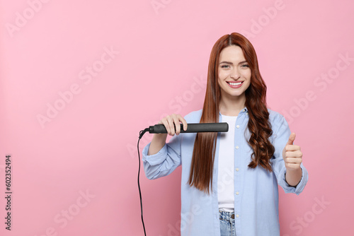 Beautiful woman with hair iron showing thumbs up on pink background, space for text