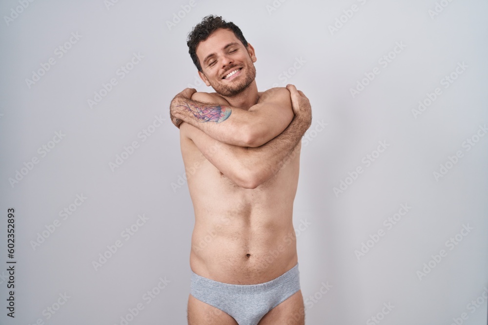Fototapeta premium Young hispanic man standing shirtless wearing underware hugging oneself happy and positive, smiling confident. self love and self care