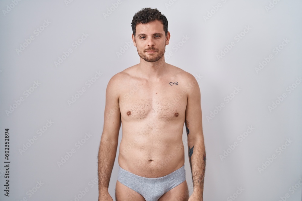 Fototapeta premium Young hispanic man standing shirtless wearing underware relaxed with serious expression on face. simple and natural looking at the camera.