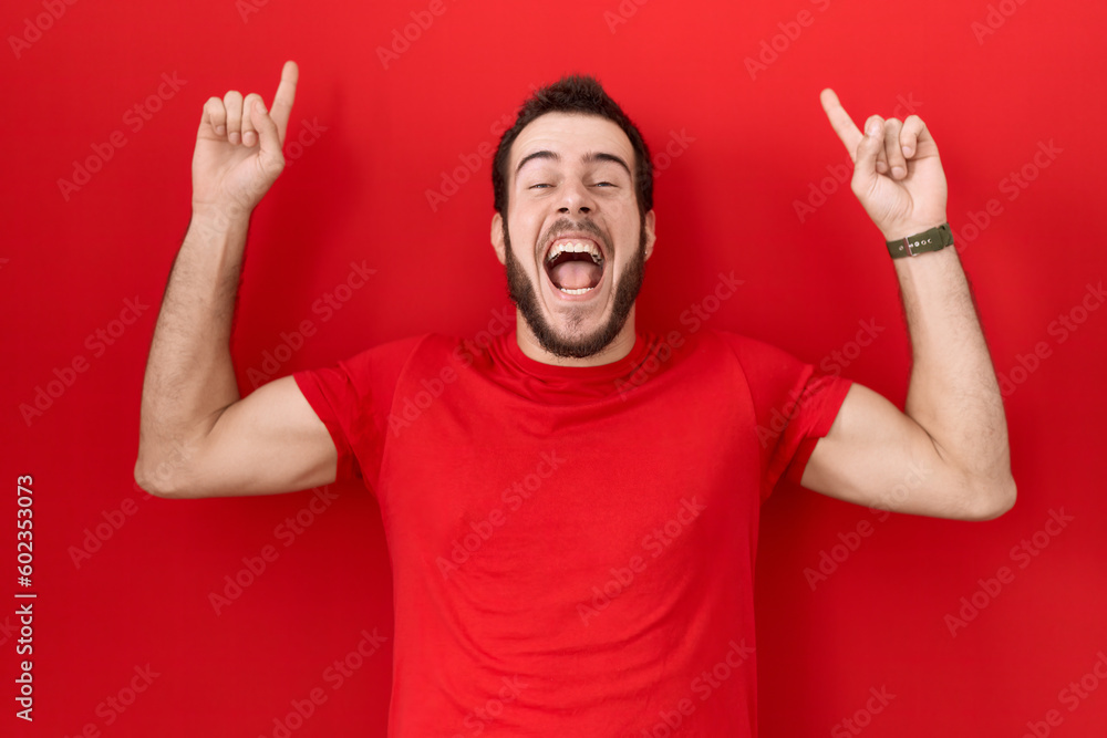 Young hispanic man wearing casual red t shirt smiling amazed and surprised and pointing up with fingers and raised arms. Young hispanic man wearing casual red t shirt smiling amazed and surprised and pointing up with fingers and raised arms.