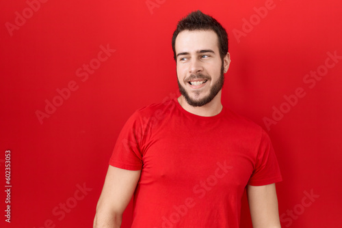 Young hispanic man wearing casual red t shirt looking away to side with smile on face, natural expression. laughing confident.