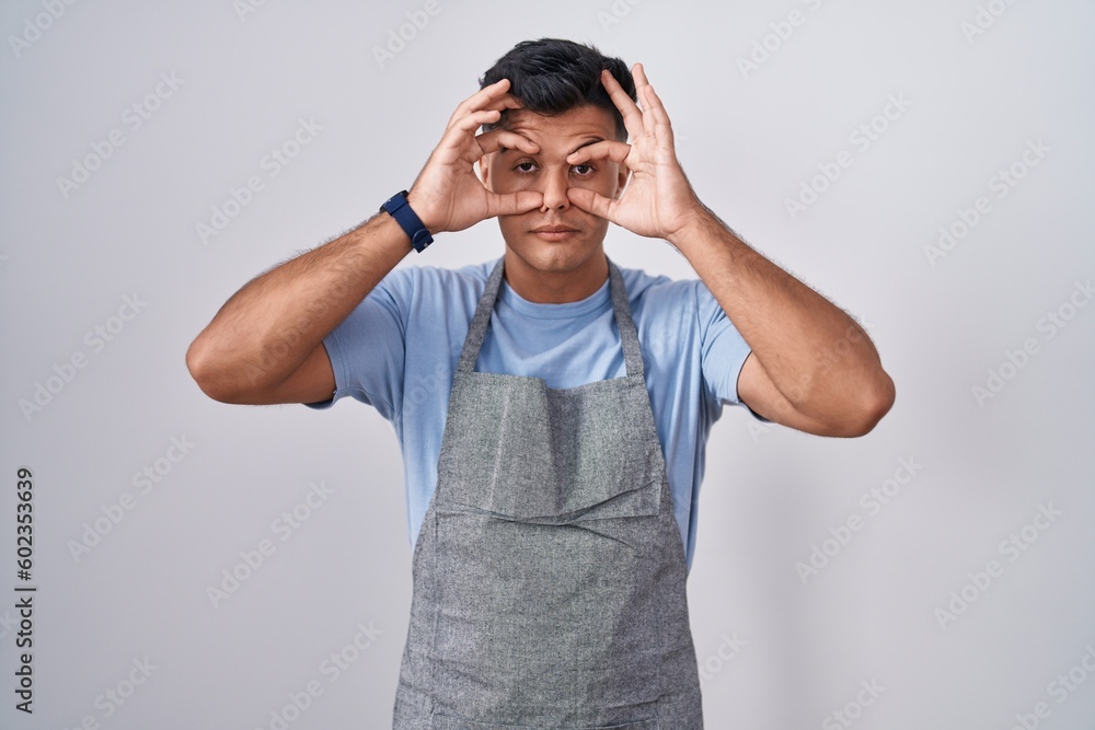 Hispanic young man wearing apron over white background trying to open eyes with fingers, sleepy and tired for morning fatigue Hispanic young man wearing apron over white background trying to open eyes with fingers, sleepy and tired for morning fatigue