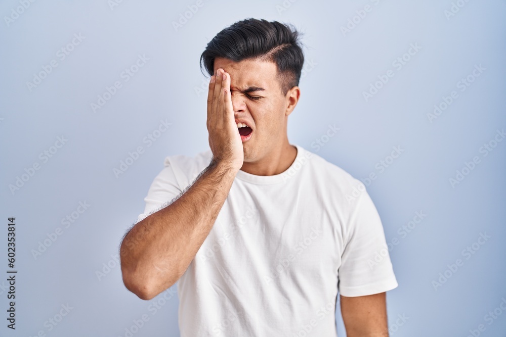 Hispanic man standing over blue background yawning tired covering half face, eye and mouth with hand. face hurts in pain. Hispanic man standing over blue background yawning tired covering half face, eye and mouth with hand. face hurts in pain.