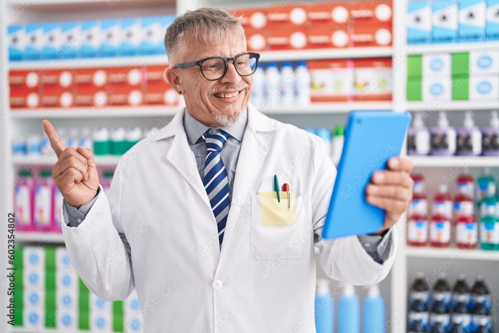 Hispanic man with grey hair working at pharmacy drugstore doing video call with tablet smiling happy pointing with hand and finger to the side Hispanic man with grey hair working at pharmacy drugstore doing video call with tablet smiling happy pointing with hand and finger to the side