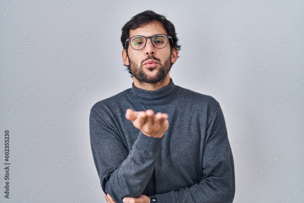 Handsome latin man standing over isolated background looking at the camera blowing a kiss with hand on air being lovely and sexy. love expression. Handsome latin man standing over isolated background looking at the camera blowing a kiss with hand on air being lovely and sexy. love expression.