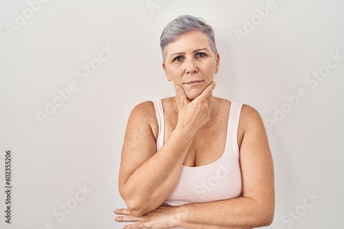 Middle age caucasian woman standing over white background looking confident at the camera smiling with crossed arms and hand raised on chin. thinking positive.