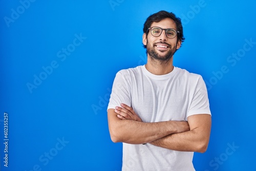 Handsome latin man standing over blue background happy face smiling with crossed arms looking at the camera. positive person.