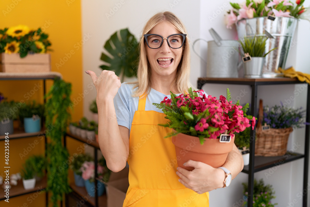 Young caucasian woman working at florist shop holding plant pointing thumb up to the side smiling happy with open mouth