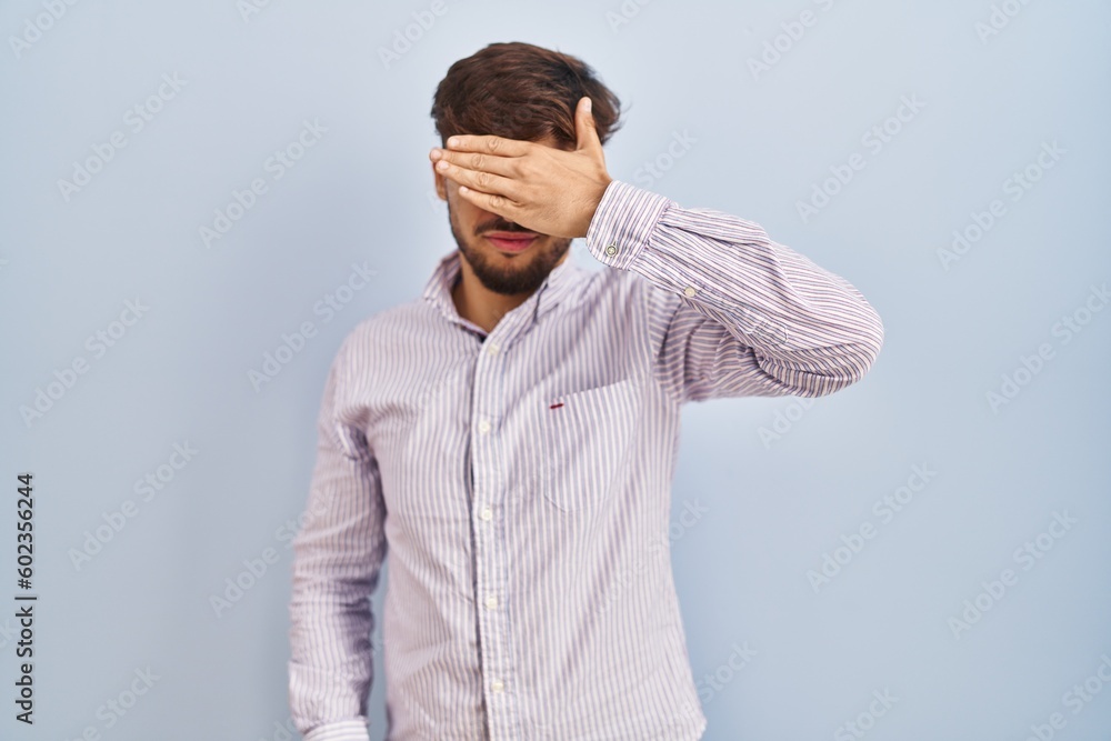 Arab man with beard standing over blue background covering eyes with hand, looking serious and sad. sightless, hiding and rejection concept