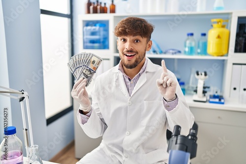 Arab man with beard working at scientist laboratory holding money surprised with an idea or question pointing finger with happy face  number one