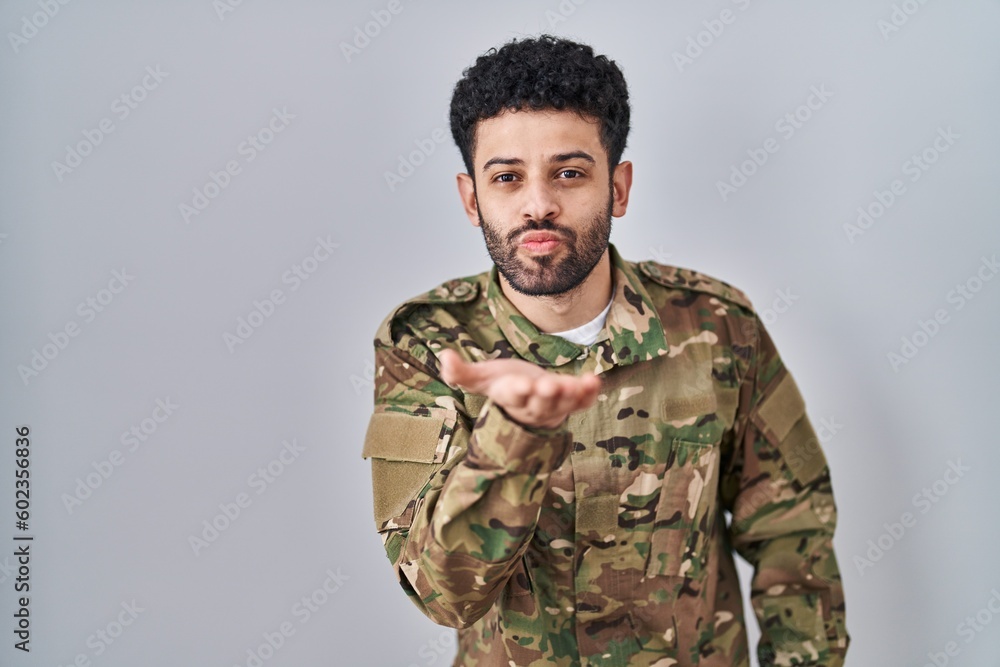 Arab man wearing camouflage army uniform looking at the camera blowing a kiss with hand on air being lovely and sexy. love expression. Arab man wearing camouflage army uniform looking at the camera blowing a kiss with hand on air being lovely and sexy. love expression.