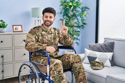 Arab man wearing camouflage army uniform sitting on wheelchair showing and pointing up with fingers number two while smiling confident and happy.